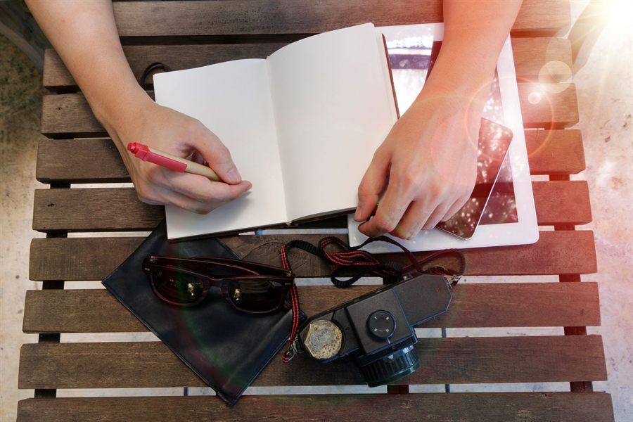 Hands writing notes in an open notebook beside sunglasses and a camera
