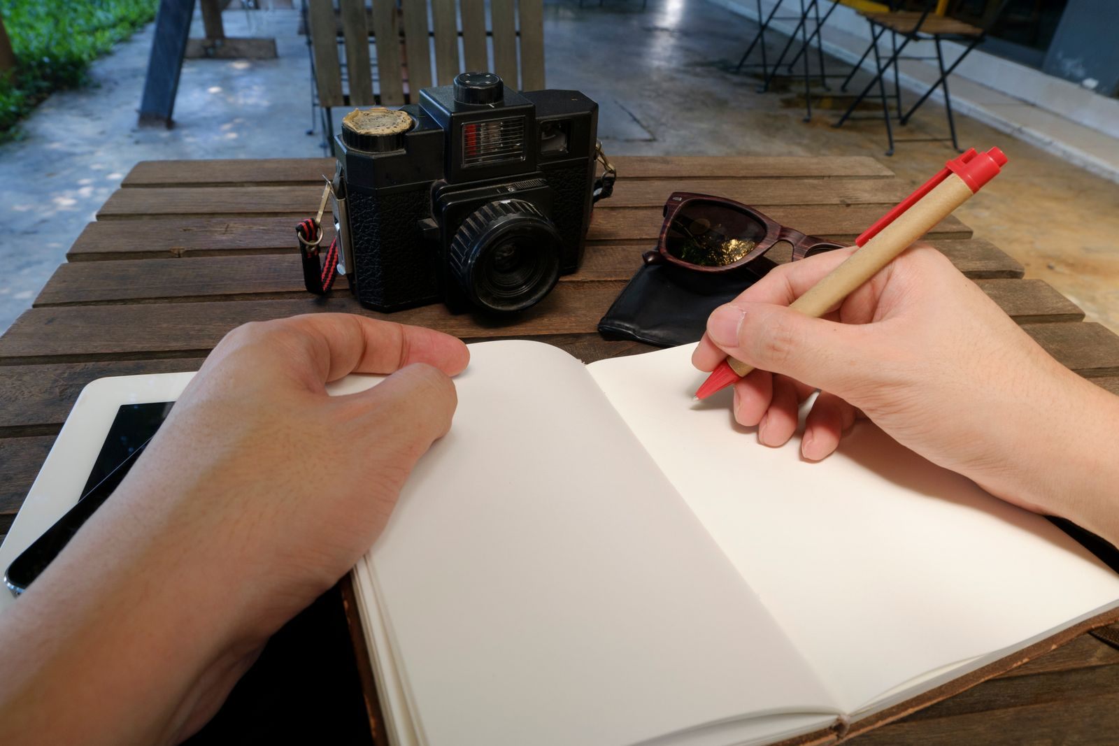 Hands writing in a notebook on a cafe table beside a camera and sunglasses