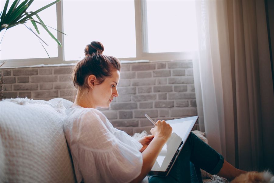 A woman on a sofa writing on a tablet by a sunny window